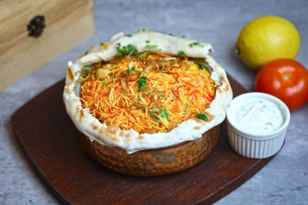 Authentic clay pot Indian biryani with raita, accompanied by lemon and tomato.